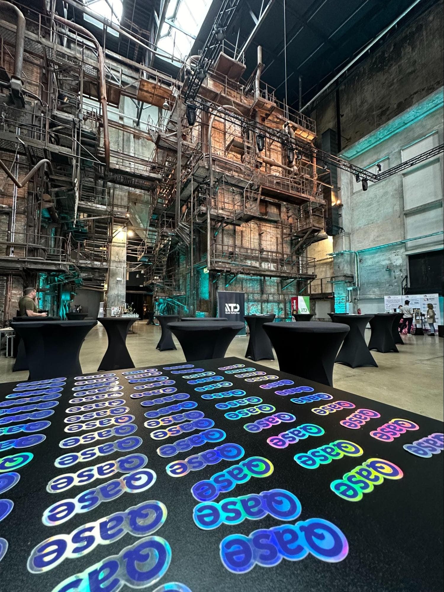 Photo with rows of Qase stickers on a table with industrial staircases and structures in the background