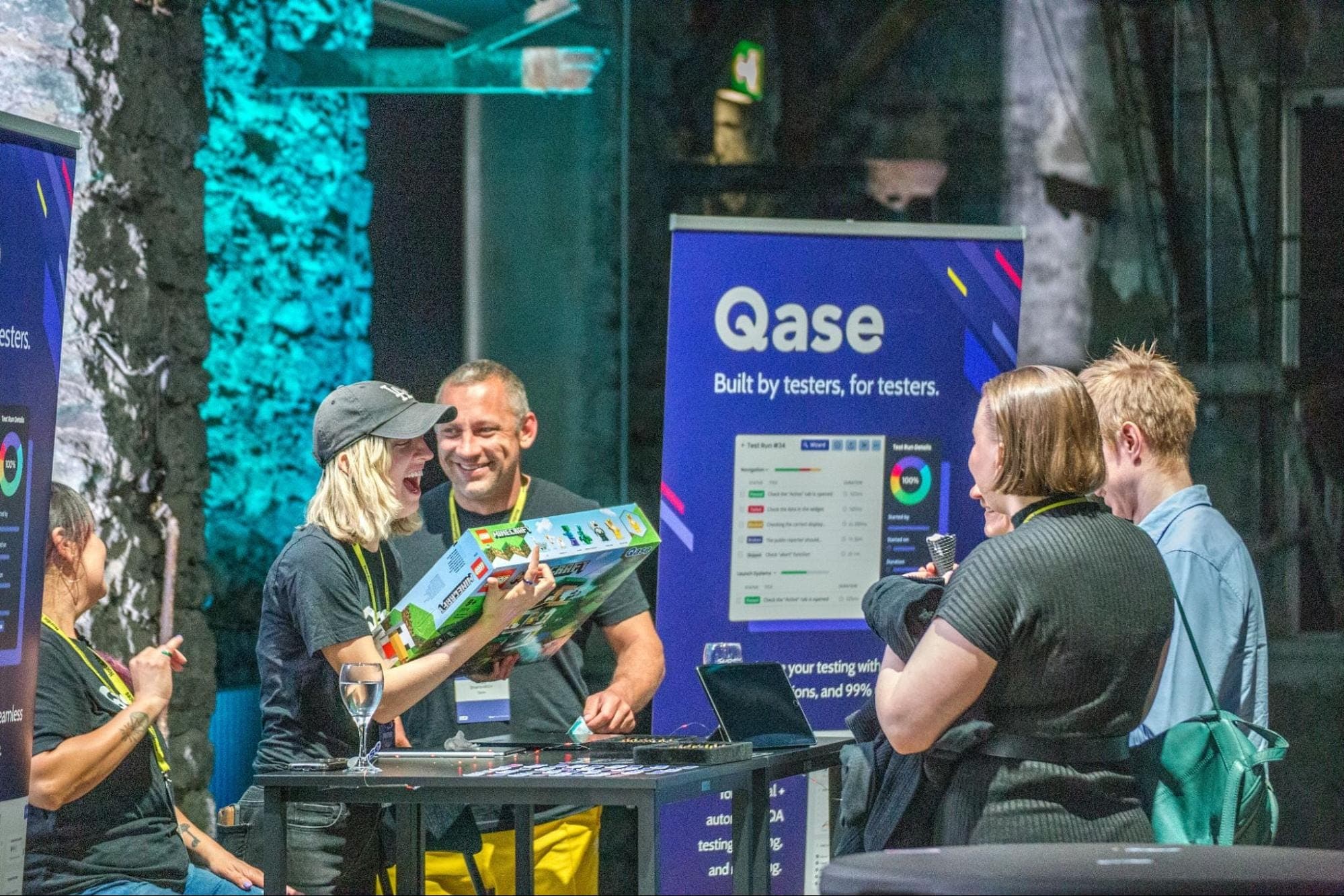 Rease, Breezy, and Vitaly laughing with Nordic Testing Days attendees at Qase Booth. Breezy is holding a Minecraft LEGO set