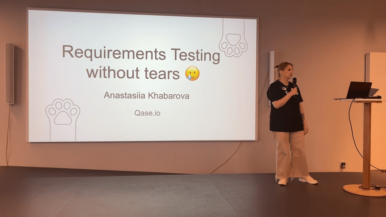 Photo of Anastasiia Khabarova wearing Qase t-shirt on stage next to slide with illustrated cat paws and text "Requirements Testing without Tears"