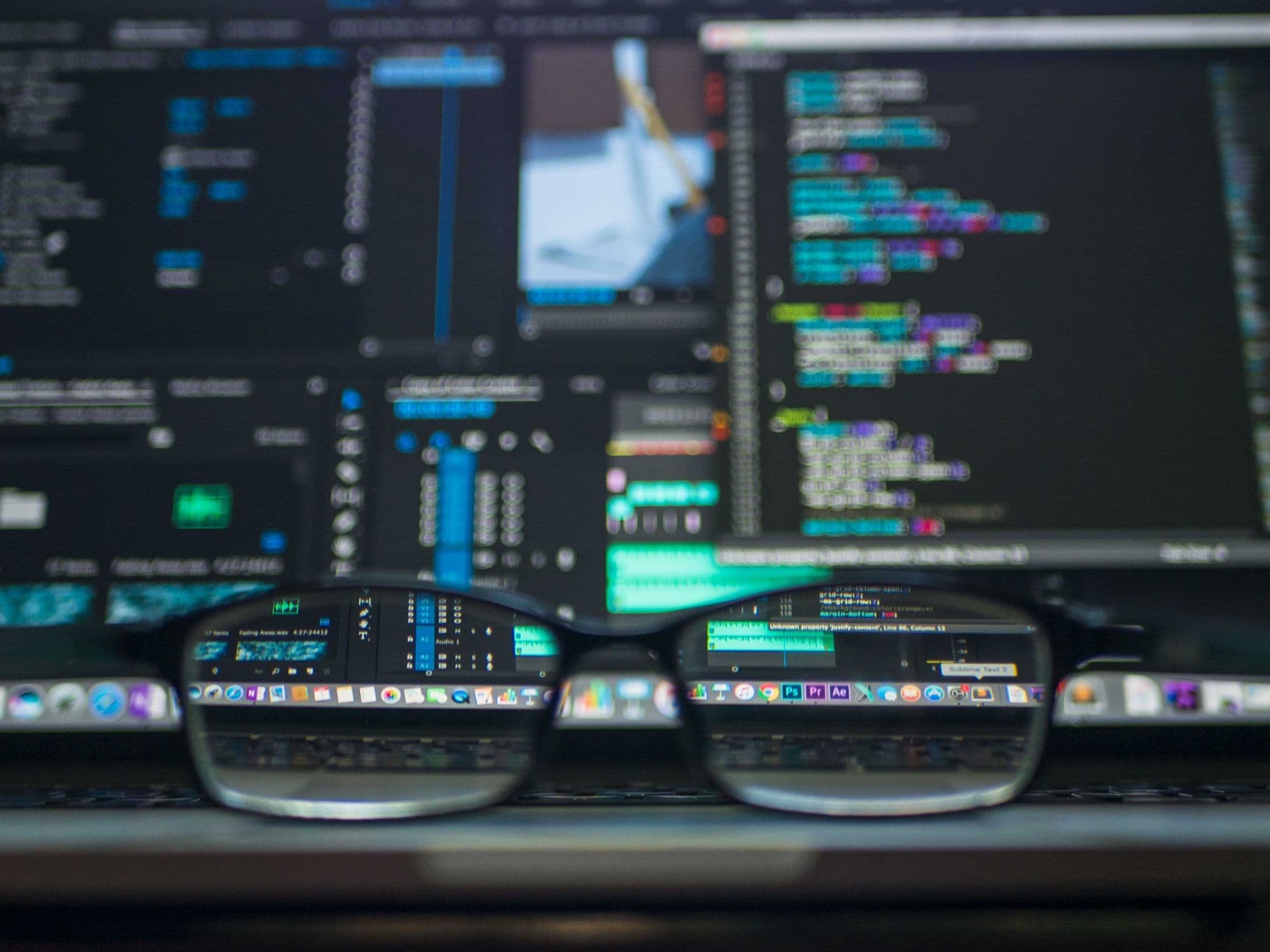 Photo of eye glasses in focus in front of a blurred computer screen showing code.